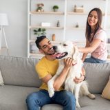 man sitting on a couch and holding a golden retriever with a woman standing behind and petting the dog